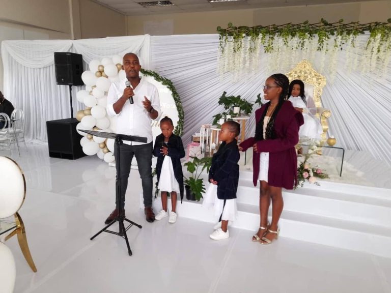 Shozi The Emcee in a white shirt speaking into a microphone at a lectern beside two young girls, with a woman seated on a gold throne in white and gold decor behind him