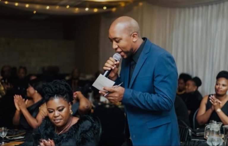 Shozi The Emcee in a blue suit holding a microphone while reading from a card at a corporate gala dinner