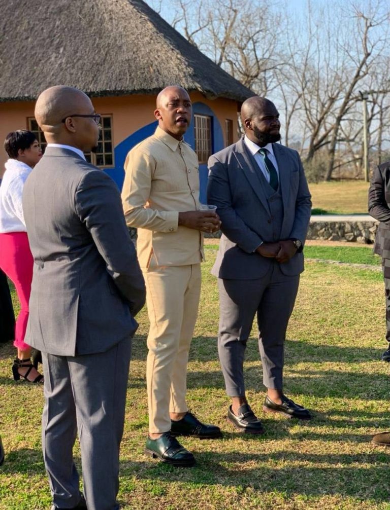 Shozi The Emcee in a cream outfit standing with two other men in suits outside a thatched roof building