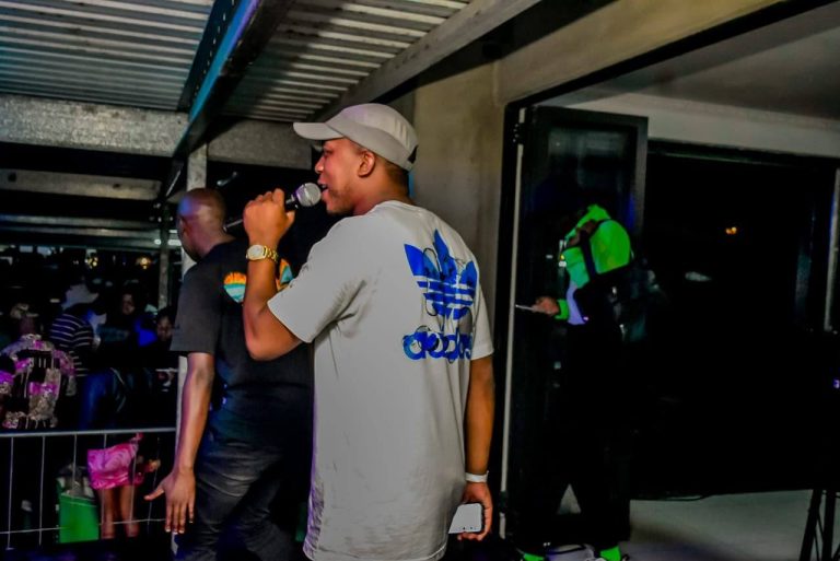 Shozi The Emcee in a grey cap and Adidas t-shirt holding a microphone side-on at a nightclub event with the crowd in the background