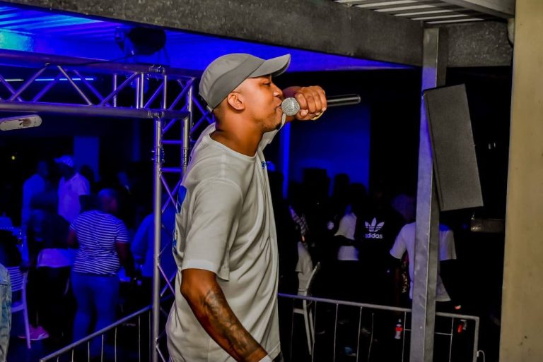Shozi The Emcee in a grey cap and t-shirt speaking into a microphone on a nightclub stage under blue lighting