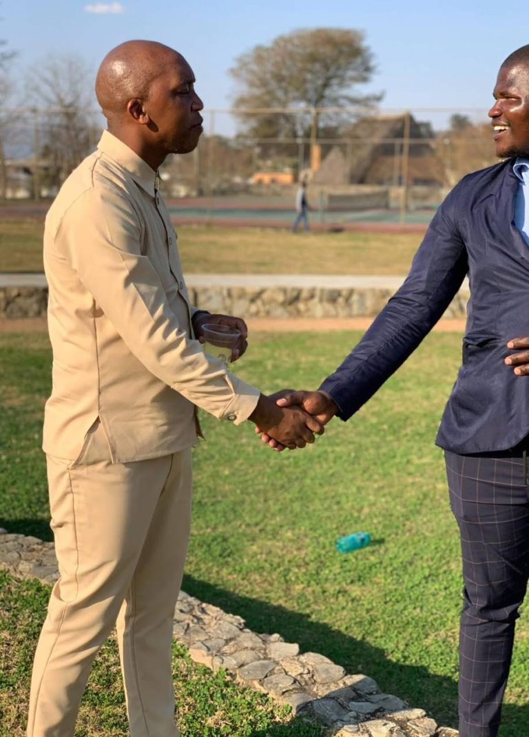 Shozi The Emcee in a beige outfit shaking hands with a man in a navy blazer on a grass lawn