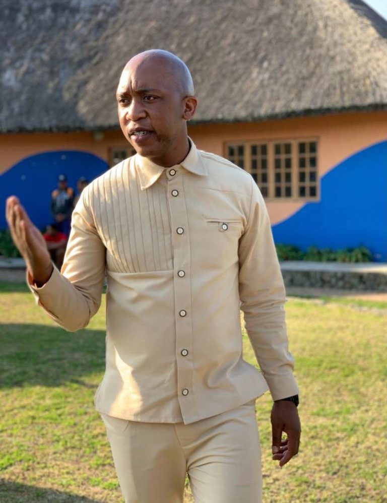 Shozi The Emcee in a cream traditional style shirt gesturing while speaking outdoors in front of a thatched roof building