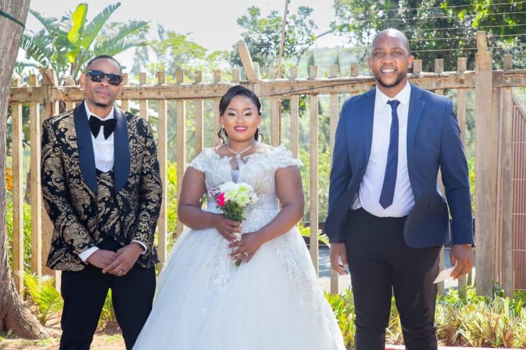 Shozi The Emcee in a black and gold patterned jacket standing beside a bride in a white gown and a groom in a navy suit outdoors