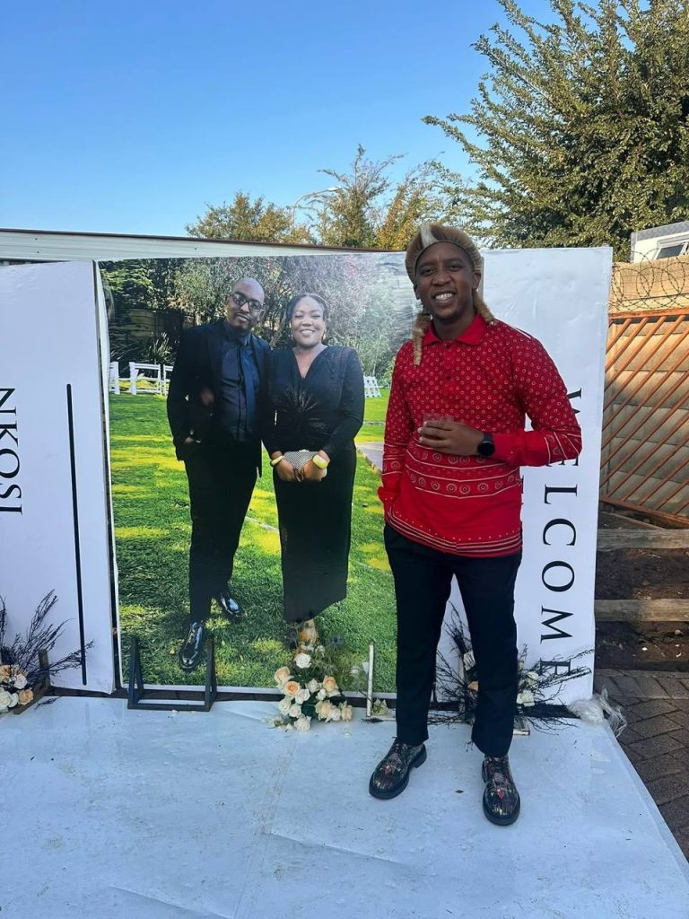 Shozi The Emcee in a red Zulu print shirt standing beside a wedding welcome board featuring the bride and groom